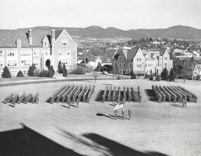 1945 photograph of Military Science Cadets. Military cadets in parade formation on campus grounds with buildings in the background.