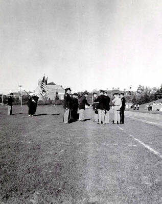 1953 photograph of Military Science Cadets. Cadet receiving an award at MacLean Field. Campus buildings visible in the background.