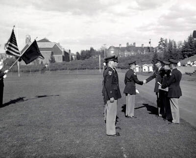 1953 photograph of Military Science Cadets. Cadet receiving an award at MacLean Field. Campus buildings visible in the background.