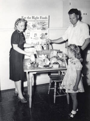 1953 photograph of Home Economics. A table with food in front of a poster that reads 'Eat the Right Foods.'