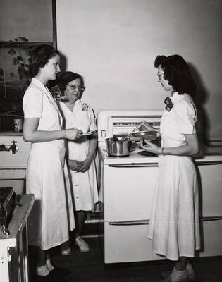 1945 photograph of Home Economics. Students gather around a stove during cooking class.