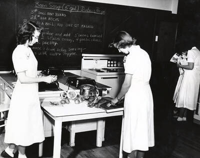 1955 photograph of Home Economics. Students cooking bean soup in a kitchen classroom. Donor: Publications Dept.