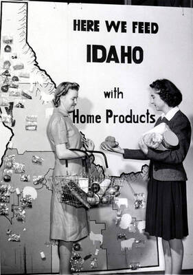 1941 photograph of Home Economics. Two students with Idaho grown products stand in front of a large poster of Idaho with the legend 'Here we feed Idaho with home products.' Donor: Publications Dept.