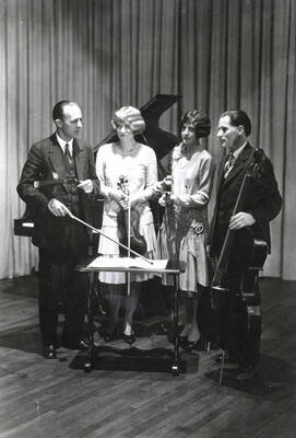 1928 photograph of Music Department. The Music Department Faculty string quartet with their instruments stand in front of a piano.
