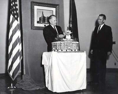1964 photograph of 75th Anniversary. Ezra M. Hawkes and James H. Roper cutting a cake shaped like the old Administration building.