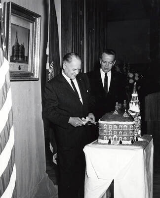 1964 photograph of 75th Anniversary. Ezra M. Hawkes and James H. Roper cutting a cake shaped like the old Administration building.