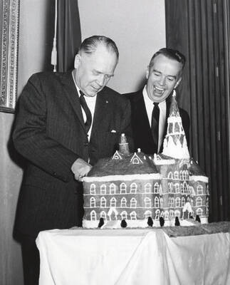 1964 photograph of 75th Anniversary. Ezra M. Hawkes and James H. Roper cutting a cake shaped like the old Administration building.