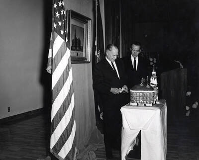 1964 photograph of 75th Anniversary. Ezra M. Hawkes and James H. Roper cutting a cake shaped like the old Administration building.