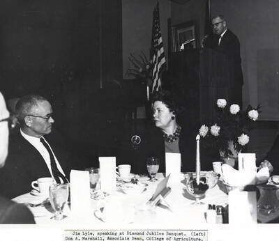 1964 photograph of 75th Anniversary. Don Marshall seated at a table with Jim Lyle standing at a lectern in the background during the Diamond Jubilee.