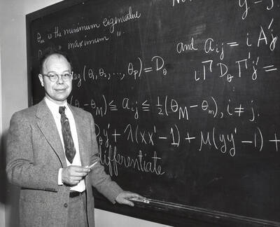 1957 photograph of Mathematics. Kenneth A. Bush in front of a blackboard.
