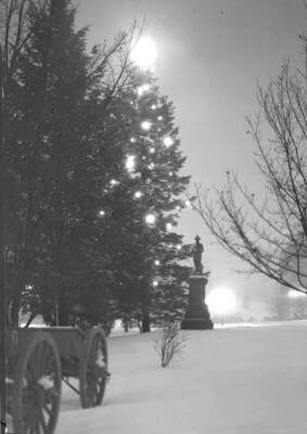 1940 photograph of the Spanish American War Memorial. Cannon in foreground, holiday lights on tree in background.
