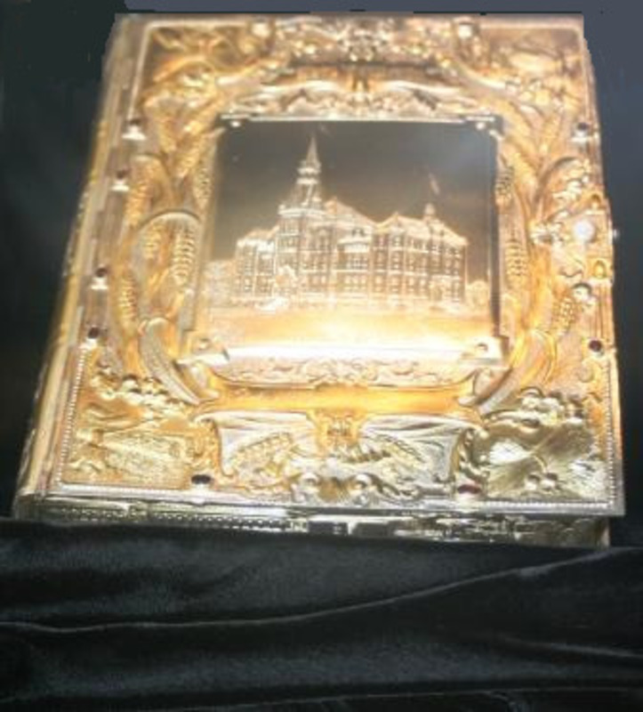 Book with an engraved image of a building and other patterns on its cover. The piece is designed by Anne Bowman and made of silver, gold, opals, and rubies.