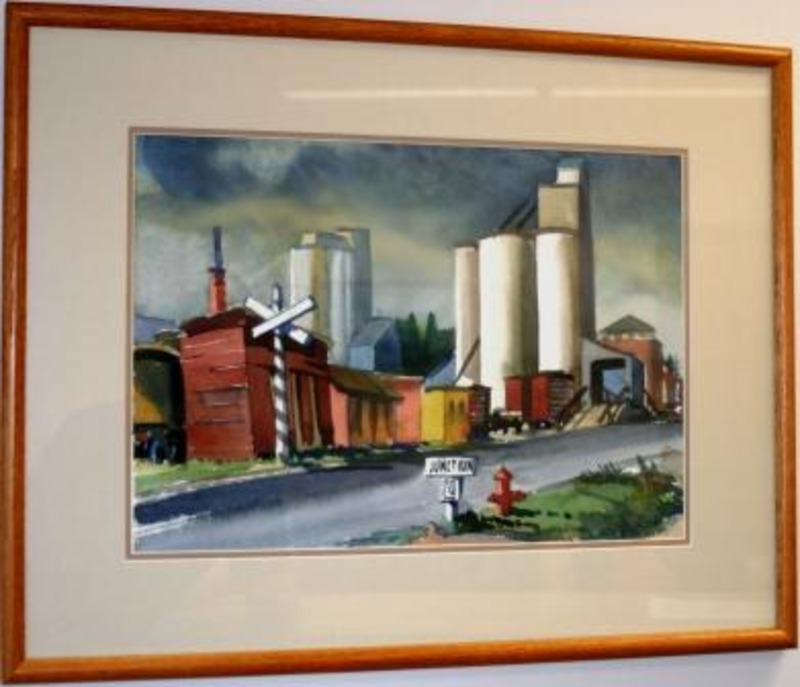 Watercolor painting depicting grain elevators by a road and a railroad. It is displayed in a light wood frame.