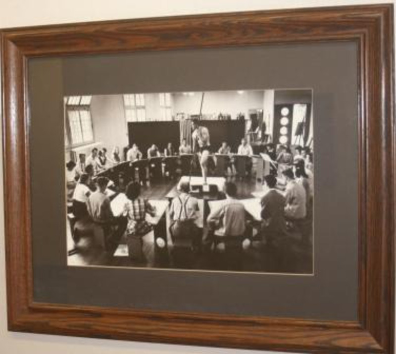 Framed black and white photograph of art class. A model poses with a stick in the middle of a circle of desks with students sketching.This piece is displayed under glass.