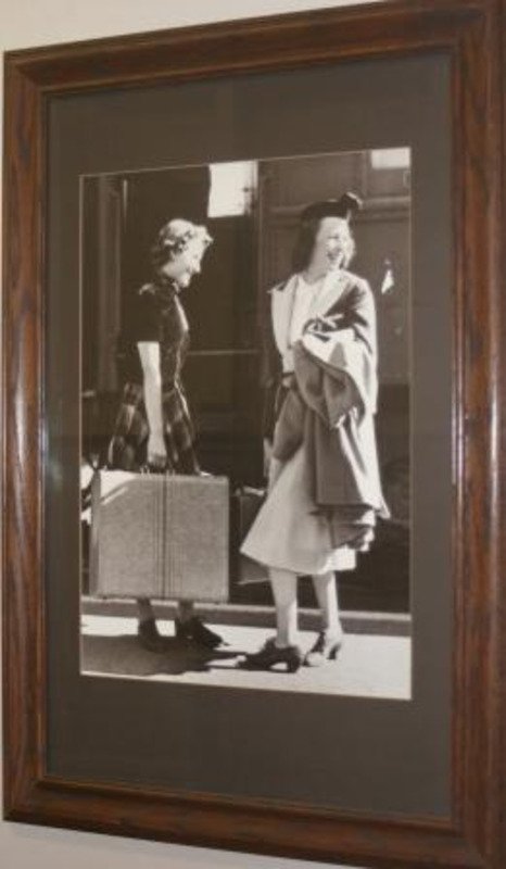 Framed black and white photograph under glass shows two female students. Both are carrying suitcases and looking towards the right in front of a train.