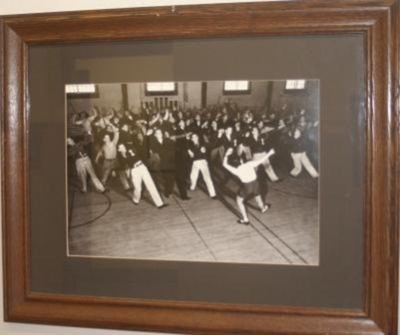 Framed black and white photograph of a men's tap class in an indoor gym. A man leads the class in front. This piece is framed under glass