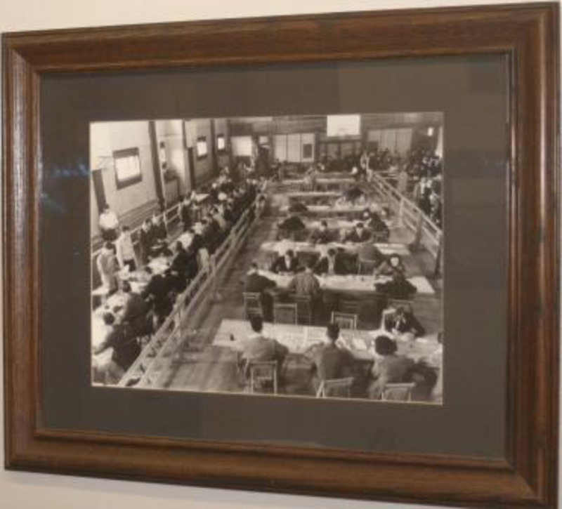 Black and white photograph of students at tables during registration. Displayed under glass in a wooden frame.