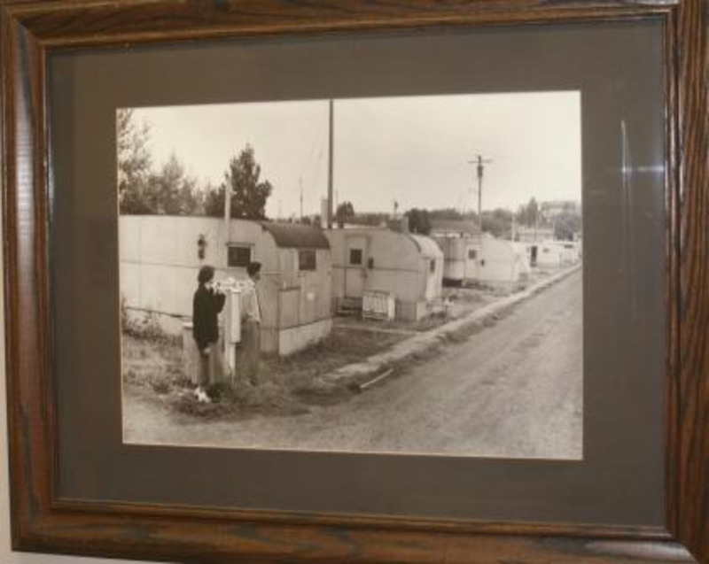 Black and white photograph of two people standing by a row of the trailers that made the Vet Village. Displayed under glass in a wooden frame.