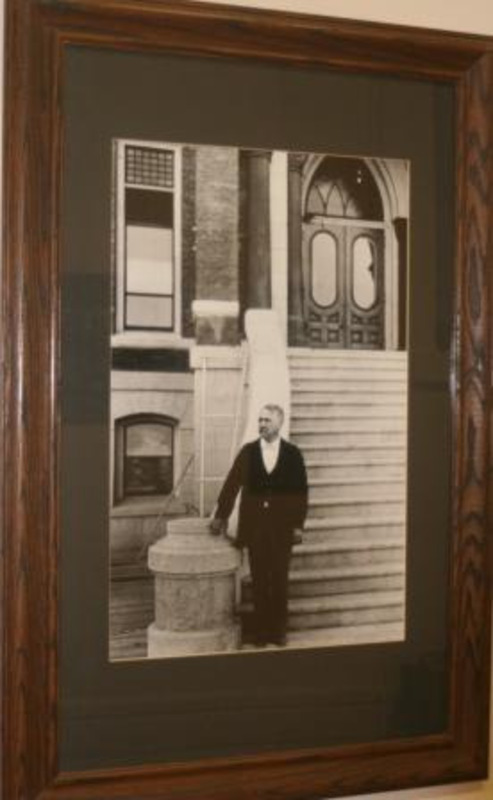 Black and white photograph of Franklin B. Gault, president of the University of Idaho 1892-1898, standing on the steps of a building on the University of Idaho campus. Displayed under glass in a wooden frame.