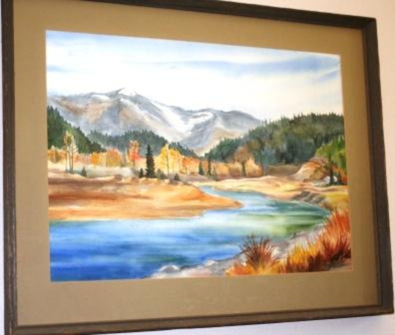Painting showing a river flowing through a forested landscape with autumnal and evergreen trees with a mountain in the background. Displayed using a light brown matte in a weathered wooden frame.