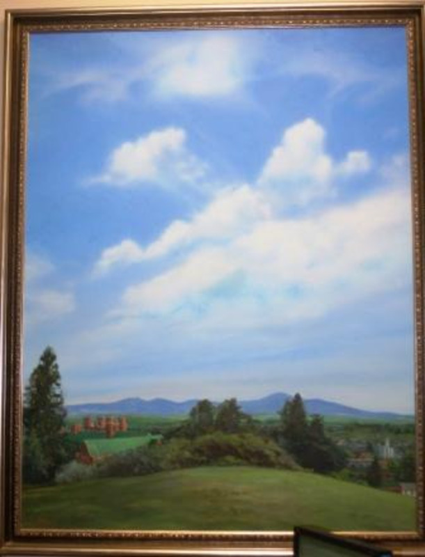 Painting showing a verdant landscape beneath a blue sky with red brick buildings, town buildings, and mountains in the background. Displayed using a decorative wood frame.