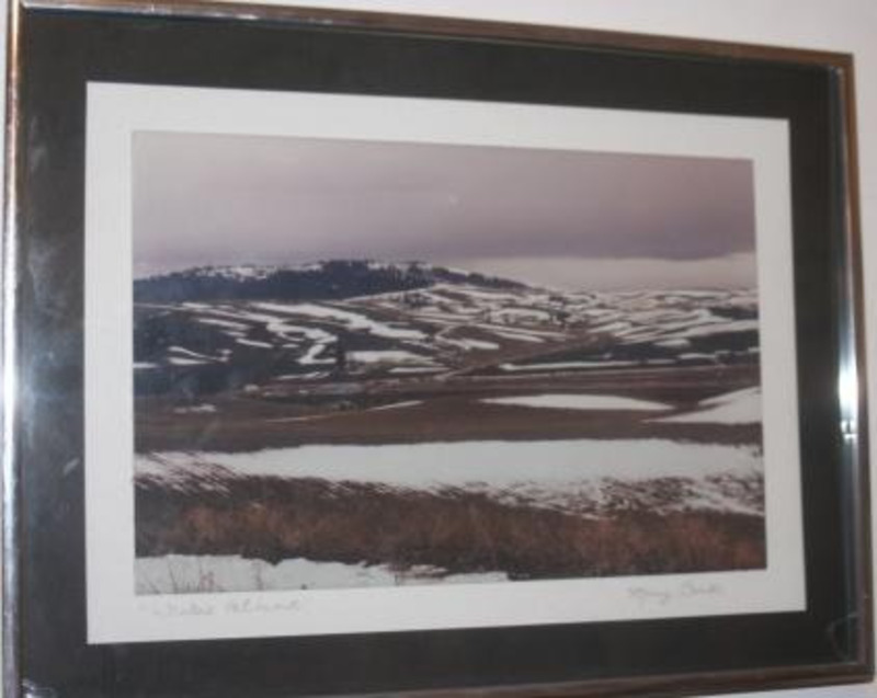 Color photograph showing a patchwork of snow on a landscape of rolling hills and distant mountains. Displayed using black and white mattes in a silver metal frame.