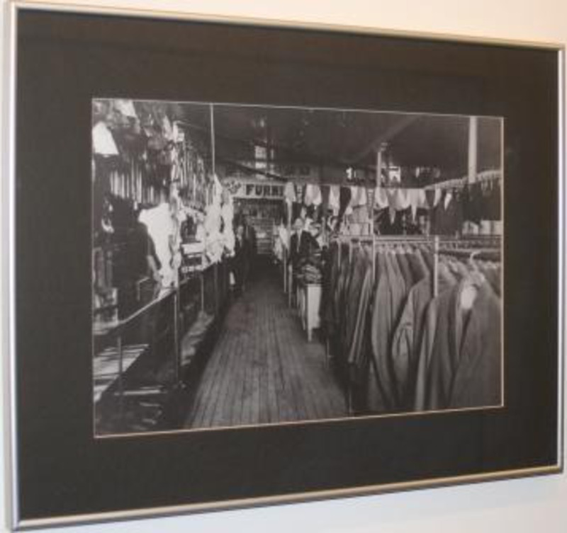 Black and white photograph showing the interior of a department store with two employees surrounded by clothing hung on racks.