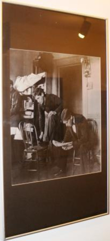 Black and white photograph showing three men in an apartment, one polishing his left shoe.