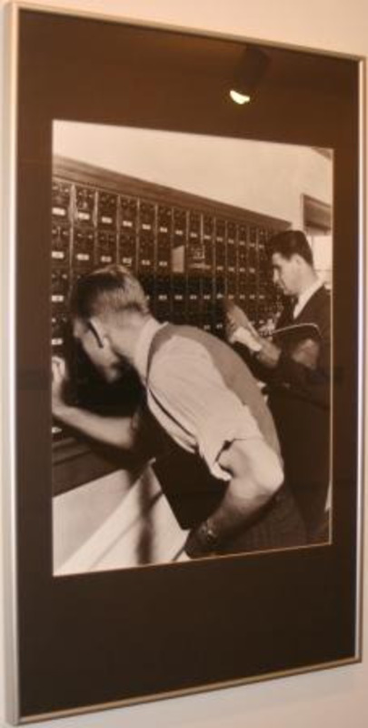 Black and white photograph showing two men getting mail from boxes on the wall.