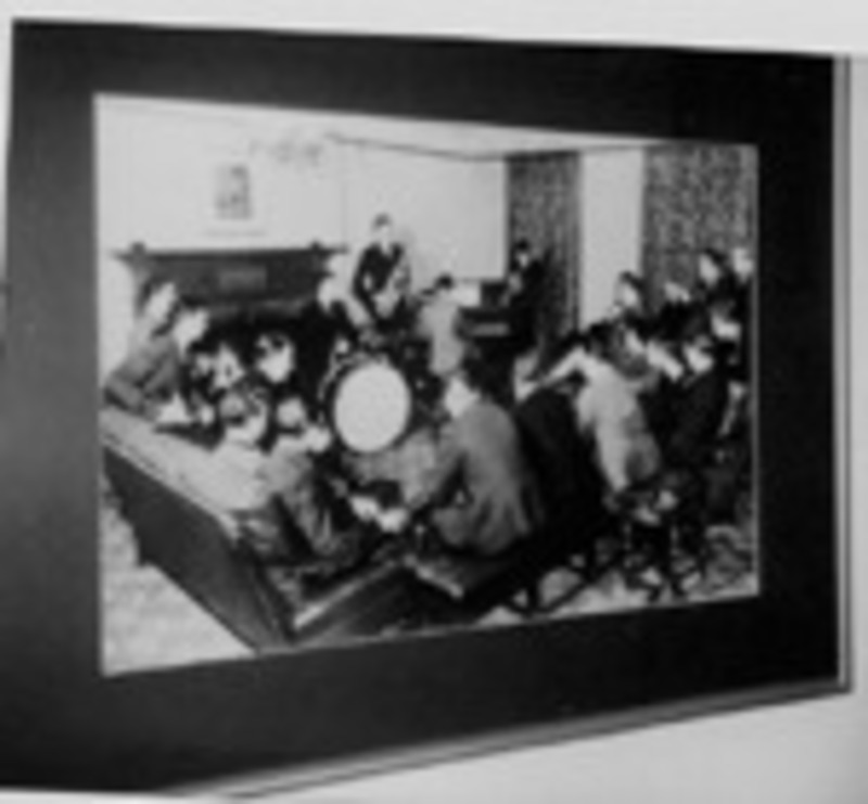 Black and white photograph of a group of men sitting in a living room around a group of people playing music.
