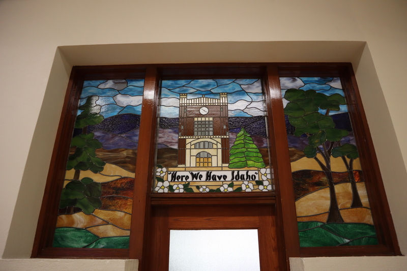 Stained glass panels depicting the University of Idaho administration building surrounded by trees, hills and mountains in the distance. Panels spelling "Here We Have Idaho" are surrounded by white flowers.