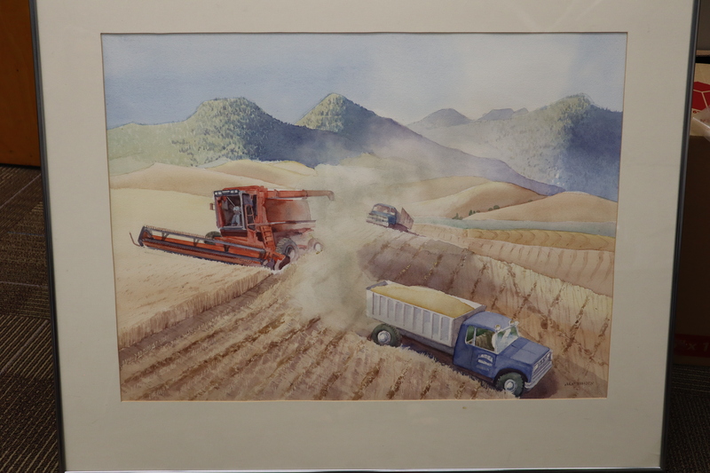 Painting of wheat being harvested on rollings hills with forested mountains in the background.