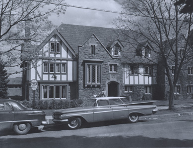 a large house with formal stone and timber design, a tidy yard, and 1950s model cars parked in front