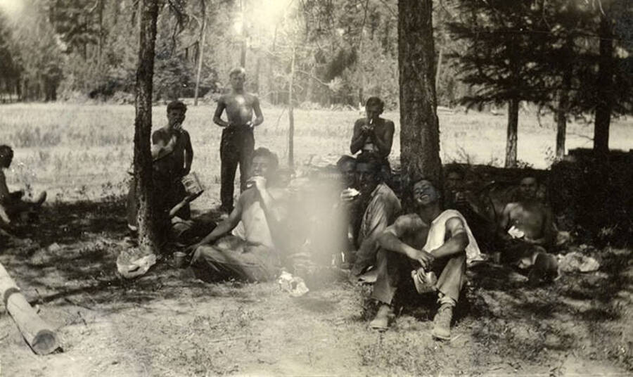 CCC work crew eating lunch in the shade. Writing below the photo reads: 'Chow'.