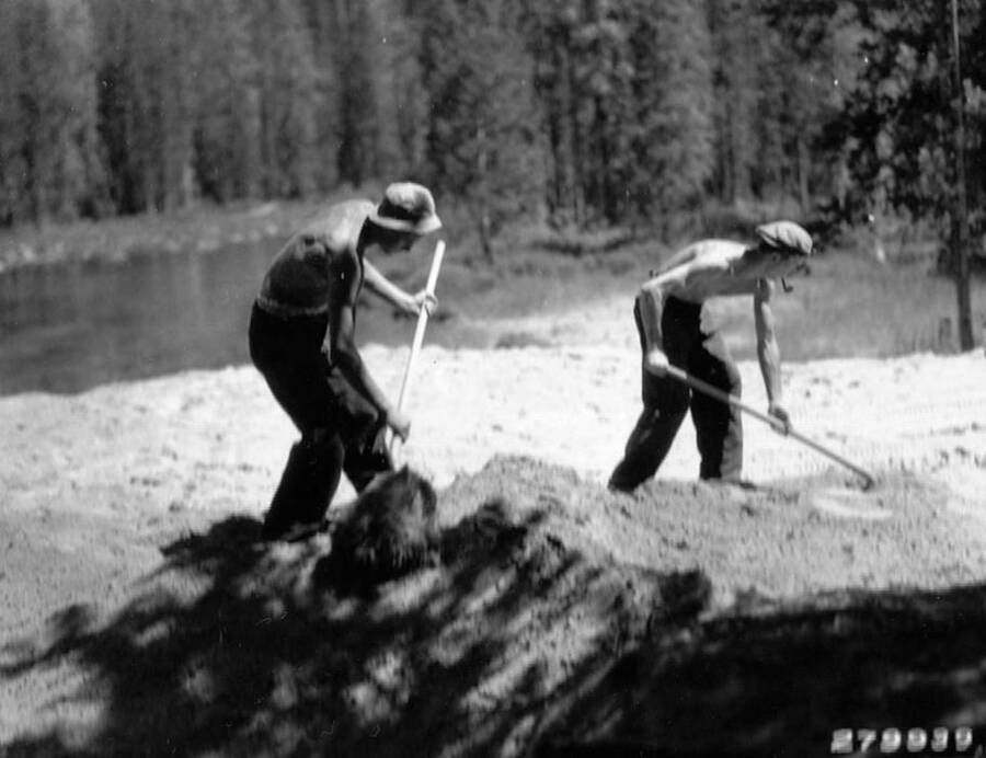 Two CCC men using shovels to spread dirt and build a road near Redfish Lake in the Sawtooth National Forest, Idaho. Note one of the men has a pipe in his mouth. Back of photo reads: 'Road work near Redfish Lake, Sawtooth National Forest, Idaho. Taken by K.D. Swan 1933.