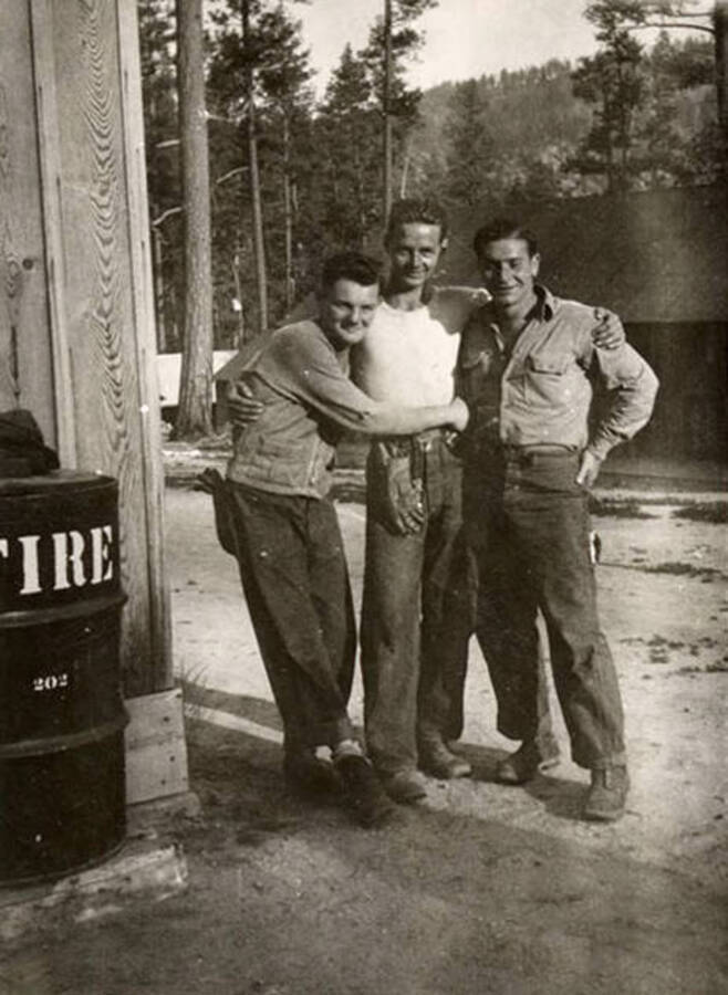 Three CCC men posed together outside a building in a CCC Camp. Next to them is a barrel reading '[T]ire 202'. Writing below the photo reads: 'Lotte - McCann -'.