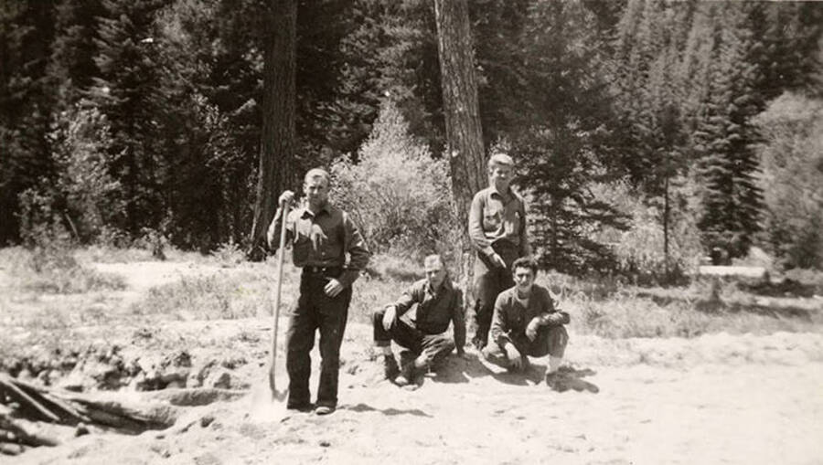 4 CCC men posing at a work site, one man is leaning on a shovel. Writing below the photo reads: 'At 'work''.