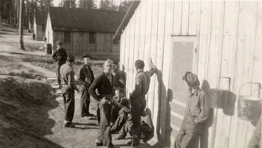 Several CCC men lounging outside of a barrack. Writing below the photo reads: 'Waiting for work call'.