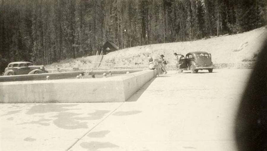 Several people relaxing at Warm Lake Swimming Pool surrounded by cars. Writing underneath each photo is a phrase that makes the sentence: 'A couple of the boys relaxing warm lake pool.' Phrase on this image is: 'A couple of'.