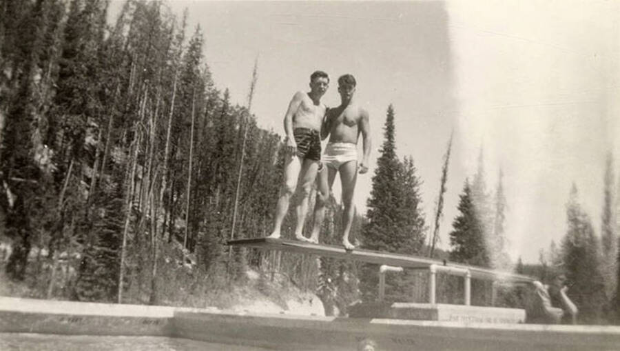 Two CCC men posing at Warm Lake Swimming Pool on the diving board. Writing underneath each photo is a phrase that makes the sentence: 'A couple of the boys relaxing warm lake pool.' Phrase on this image is: 'relaxing in'.