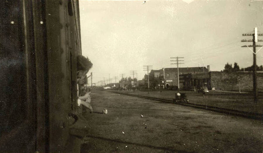 CCC men stick their heads out of the windows of a moving train to gain a view of the small town they are passing. Another set of railroad tracks can be seen, as well as a row of power lines and several buildings.