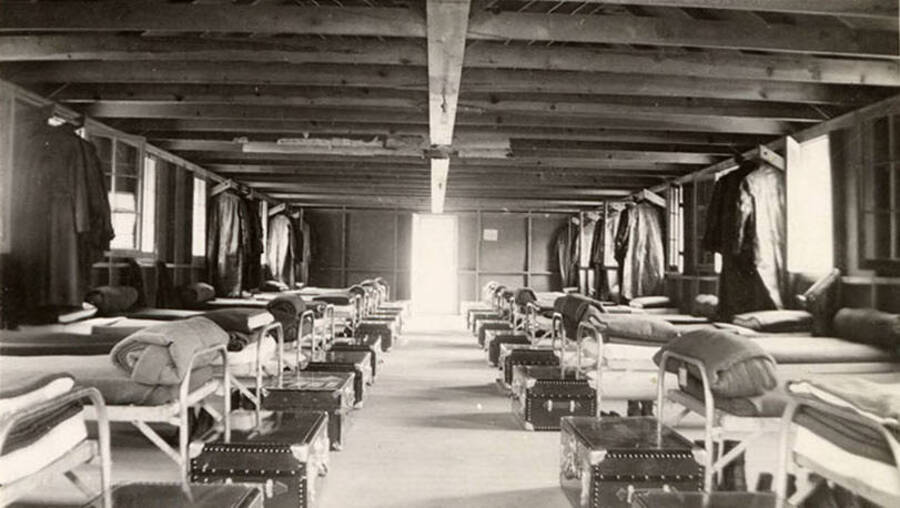 Interior view of barrack #5 in CCC Camp F-168. Beds are neatly made and trucks sit at the end of each one. Coats hang in pairs along the row of beds against the wall. Writing under the photo reads: 'Barracks #5'.