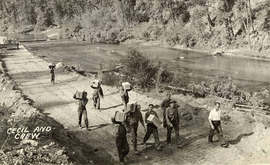 Photo of a CCC work crew walking along a road with their supervisor. Some of the men are carrying boxes. A bulldozer can be seen working on the road in the background. The river runs by the river. Writing on the photo reads: 'Cecil and Crew'.