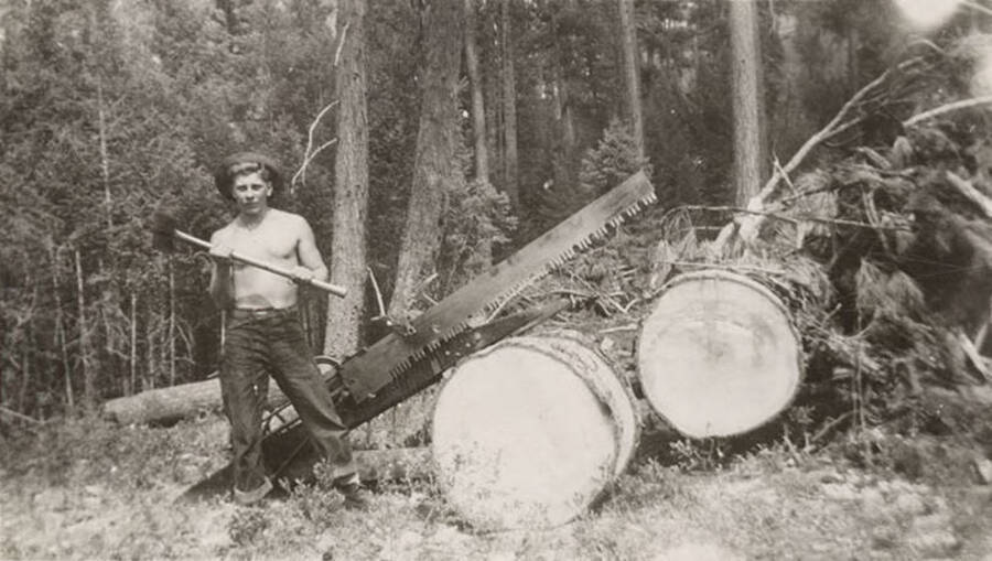 Photo of a CCC man posing with an axe next to a dragsaw and two felled trees. Writing under the photo reads: 'Bubby - 'Working''.