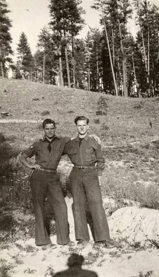 Two CCC men posing for a photo with a wooded hill in the background. In the foreground the photographer's shadow can be seen falling across the ground.