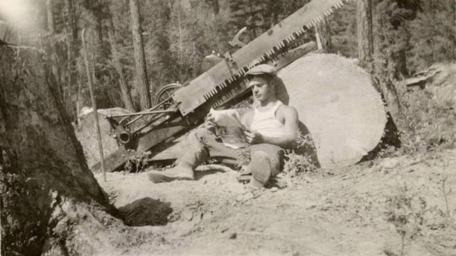 Photo of a CCC man leaning against a felled tree and reading while sitting near a dragsaw. The stump of the tree is in the foreground. Writing under the photo reads: 'Caught in the act'.