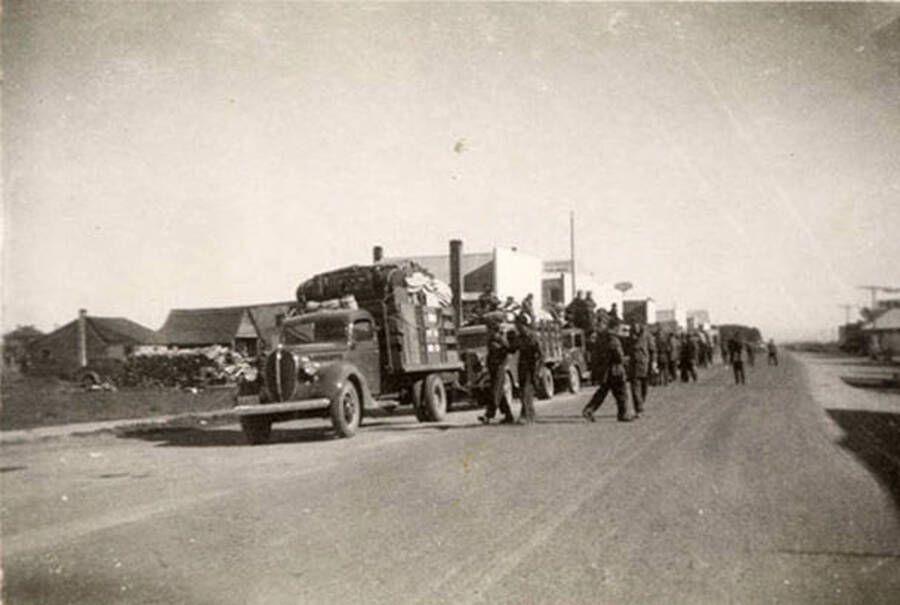 Crew of CCC Men and a convoy of trucks on a paved road with a town in the background. Writing under the photo reads: 'on the march'.