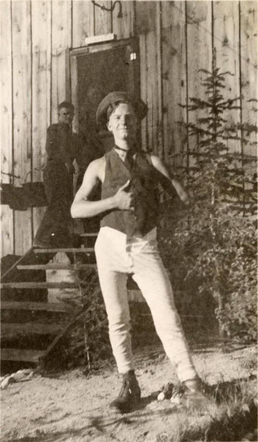 Photo of a CCC man posed in front of his barrack, another man can be seen on the steps of the barrack. Writing under the photo reads: 'Heinson'.