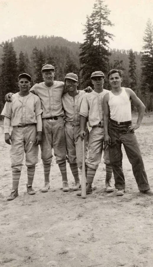 Five CCC men posing on a baseball field with wooded hills in the background. One of the players is holding a bat.