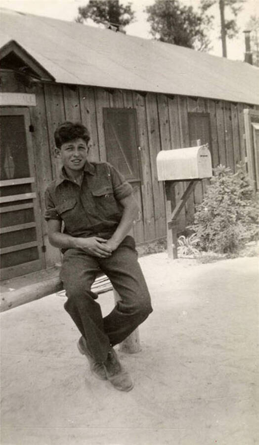 Photo of a CCC man sitting outside of a camp building with a mailbox in front of it. Writing under the photo reads: 'Lotte'.